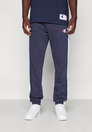 Pantalons de jogging bleu marine pour hommes avec des bandes blanches sur les côtés, des poignets élastiques et une taille élastique. Le tissu semble doux, avec un petit logo sur la cuisse gauche.