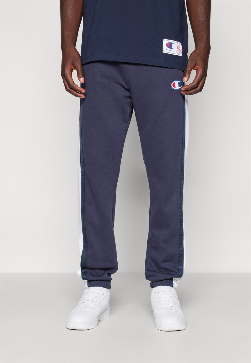 Pantalones jogger para hombre en color azul marino con franjas laterales blancas, puños elásticos y cintura elástica. La tela parece suave y presenta un pequeño logo en el muslo izquierdo.