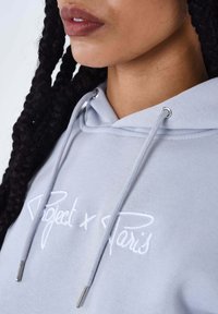 Hoodie in lichtgrijs met trekkoorden en zilveren metalen accenten. Voorzien van een geborduurd "Project x Paris" logo in het wit.