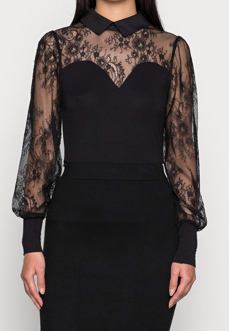 Haut noir à manches longues avec un corsage en dentelle transparente, col classique et matière noire ajustée à la taille. La dentelle présente des motifs floraux.