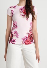 Camiseta de manga corta floral en rosa claro con ricos patrones de rosa y orquídeas. Tejido suave, diseño ajustado, escote redondeado, combinada con pantalones blancos.