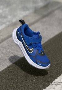 Blå idrettssko med mesh-overdel, hvit såle, svarte detaljer, snøring og borrelåslukking. Har Nike-logo i hvitt på siden.