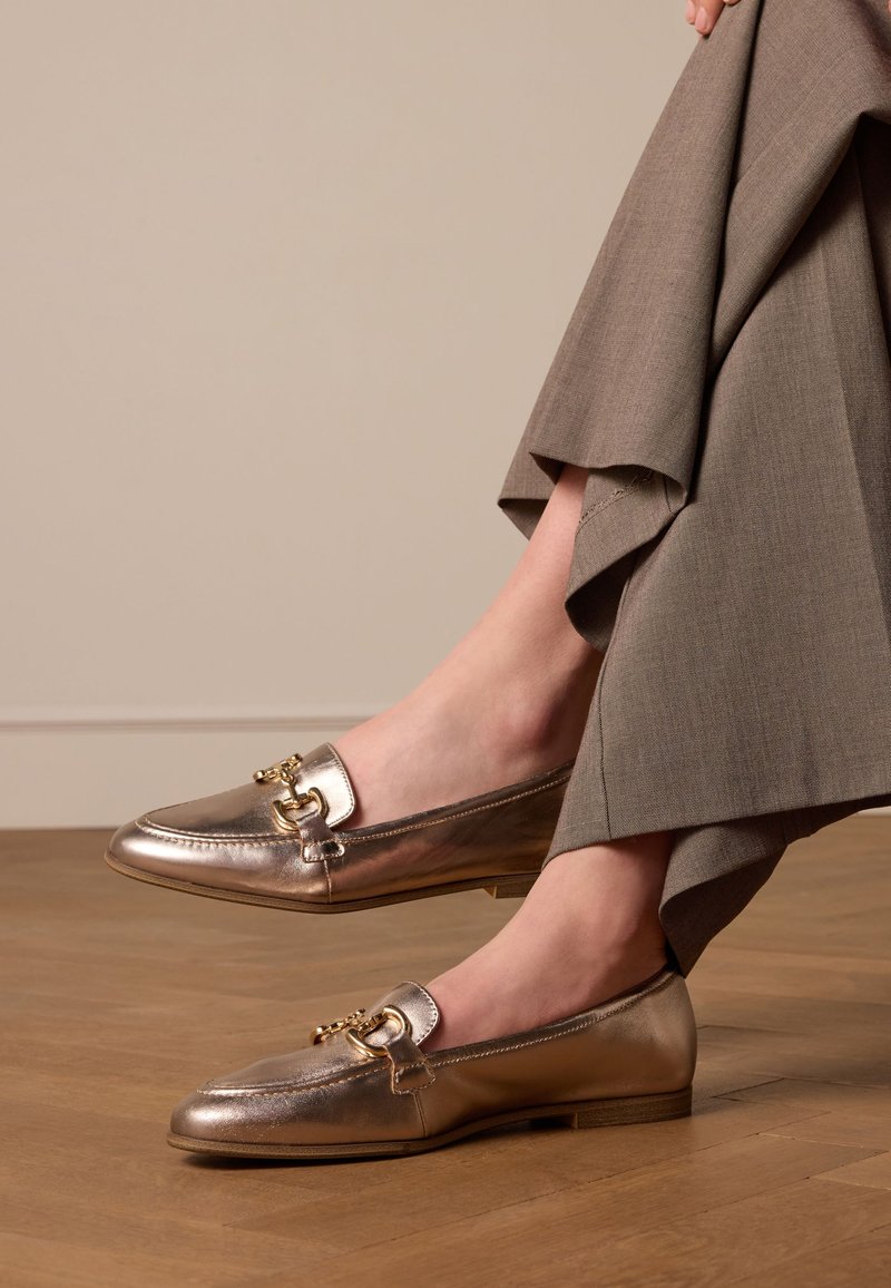 Mocassini metallici dorati con punta tonda, caratterizzati da un elegante accento di catena dorata. Parte superiore in pelle texturizzata e suola piatta in legno.