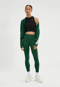 Leggings deportivos verdes y un top corto negro con una chaqueta verde a juego, de frente abierto. Rayas blancas en las mangas, textura de tela suave.