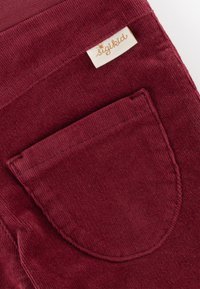 Burgunderfarbene Cordstoff mit einer großen Rückentasche und einem eingenähten Etikett mit goldfarbener Schrift. Glatte Textur mit vertikaler Rippung.