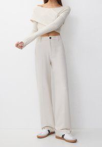 Vrouw in een off-white geribbelde gebreide crop top, high-waisted wijdvallende crèmekleurige broek en witte sneakers met zwarte strepen.