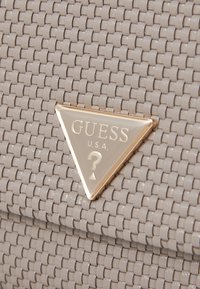 Šedá pletená kabelka s trojúhelníkovou zlatou destičkou s logem "GUESS" a detailem otazníku, zvýrazňující její texturovaný materiál.