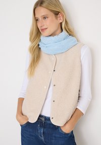 Chaleco de forro polar beige con botones a presión, llevado sobre una camisa de manga larga blanca. Bufanda azul suave colgada alrededor del cuello, complementada con jeans oscuros.