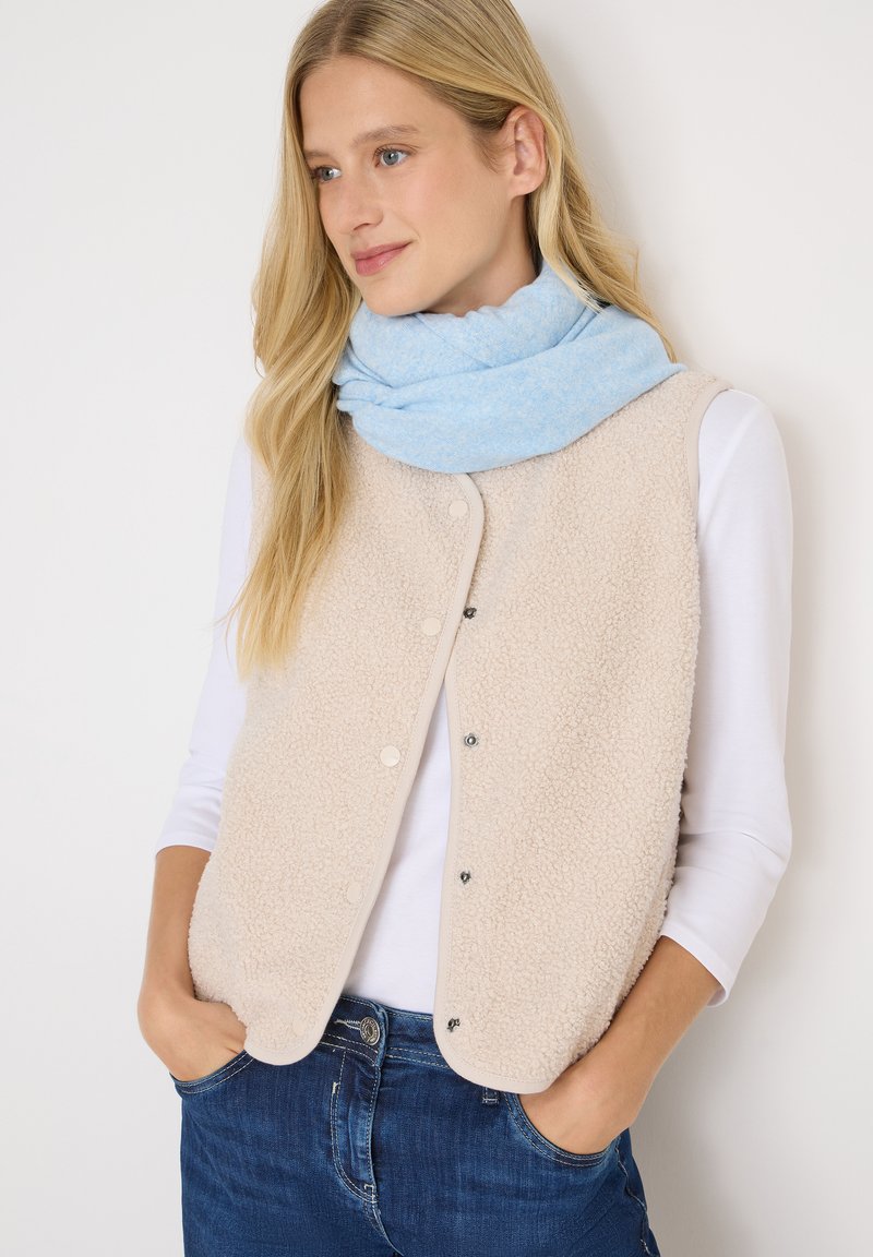 Chaleco de forro polar beige con botones a presión, llevado sobre una camisa de manga larga blanca. Bufanda azul suave colgada alrededor del cuello, complementada con jeans oscuros.