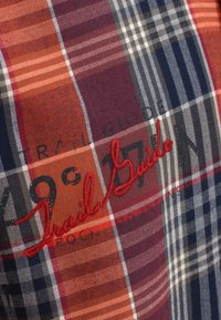 Il tessuto a quadri presenta colori blu navy, rosso, arancione e bianco con una superficie testurizzata. Il design include testo ricamato in rosso in grassetto.