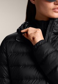 Črna puhovka z nagubanimi paneli, stoječim ovratnikom in zadrgo. Model prilagaja tekstilno zanko na ovratniku.