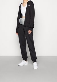 Svart zip-up huvtröja, vit crop top, svarta sweatpants, silverfärgad bältesväska, vita sneakers. Mjukt tyg, avslappnad passform, huva och ribbade manschetter.