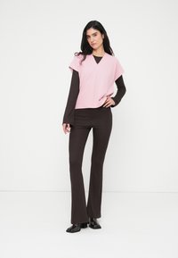 Ανοιχτό ροζ τοπ με φαρδιά μανίκια πάνω από καφέ μακρυμάνικο πουκάμισο, συνδυασμένο με στενά καφέ καμπάνα παντελόνια. Λαμπερά μαύρα παπούτσια με τακούνι.