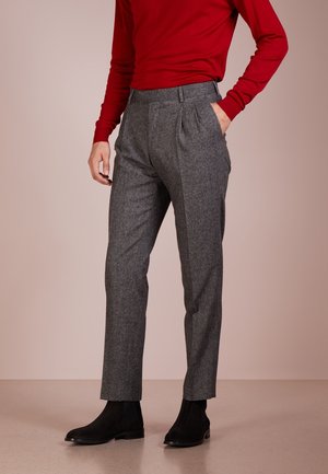 Personne portant un pantalon gris tailleur avec des plis, un haut rouge à manches longues et des bottines noires, debout avec une main dans la poche.