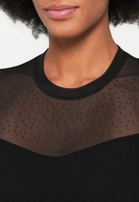 Melns tops ar caurspīdīgu acs paneli, kurā ir punktiņu tekstūra, ribota auduma apakšdaļa un apaļa kakla izgriezums.