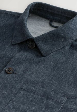 Camisa de mezclilla oscura con cuello puntiagudo, dos botones negros y un bolsillo en el pecho. La tela tiene una textura sutil y un acabado impecable.