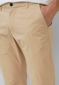 Mann in beigen Hosen mit einem schwarzen Knopf, die Hand in der rechten Tasche vor einem grauen Hintergrund platziert.