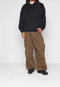 Svart fleecehuva med ficka fram, kombinerad med lösa, beige cargobyxor med sidfickor och snörning i midjan. Svarta sneakers.