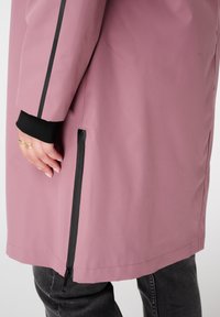 Close-up van een persoon die een mauve jas draagt met zwarte rits en streepdetails, zwarte manchetten, gouden ringen en donkergrijze jeans.