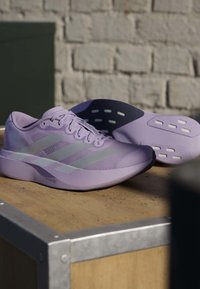 Para jasnofioletowych butów do biegania Adidas z białymi paskami, spoczywająca na drewnianym pudełku na tle ceglanej ściany.