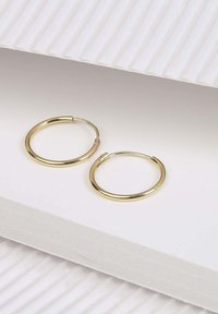 Boucles d'oreilles en or en forme de créole avec une surface lisse et réfléchissante, présentant un design simple et un fermoir sécurisé, exposées sur un papier blanc texturé.