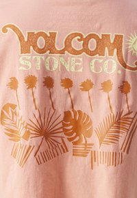 Rožinė medvilninė marškinė su dideliu oranžiniu grafinio dizaino, kuriame pavaizduoti tropiniai augalai, ir stilizuotu užrašu "Volcom Stone Co.".