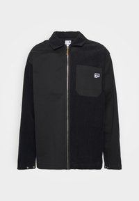 Veste noire en matériaux mélangés, dotée d'une fermeture éclair, d'un col, d'une poche sur la poitrine gauche et d'une texture en corduroy contrastante d'un côté.