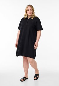 Zwarte polojurk met korte mouwen, knopen bij de kraag en een ontspannen pasvorm. Gecombineerd met zwarte slidersandalen met decoratieve accenten.