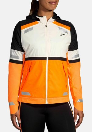 Frau trägt eine Kapuzenjacke in Weiß, Orange und Schwarz mit reflektierenden Streifen an Ärmeln, Brust und Taille, vorne zugezippt.