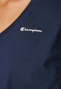 Navy zila kokvilnas T-kreklu ar V veida kakla izgriezumu un baltu izšūtu "Champion" logotipu kreisajā augšējā stūrī. Gluda tekstūra.