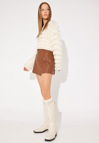 Mini gonna in pelle marrone con occhielli metallici, maglione bianco testurizzato con frange, e stivali da cowboy bianchi fino al ginocchio con suole nere.