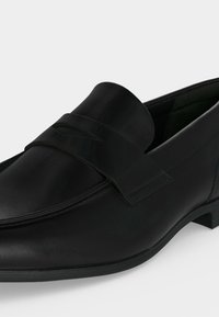 Mocassin homme en cuir noir sans lacets avec un petit talon et un détail de bride sur le dessus.