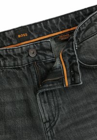 Vaqueros de mezclilla negra con un corte regular. Presentan una bragueta con cremallera, costuras marrones y una etiqueta BOSS en la cintura. Tejido con textura.