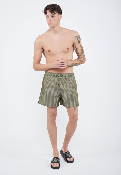 Νεαρός άνδρας χωρίς πουκάμισο με τατουάζ στο δεξί χέρι και πόδι, φοράει πράσινα ριγέ σορτς και μαύρα σανδάλια με φάσα σε λευκό φόντο.