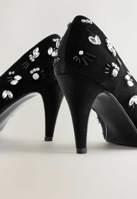 Pantofi negri cu toc înalt, realizați dintr-un material texturat, împodobiți cu cristale clare și negre în diverse forme. Design cu vârf rotund.