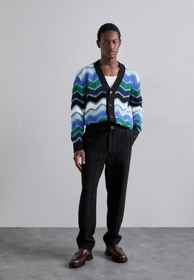 Knitted cardigan with multicolored wavy patterns in blue, green, and black, featuring a black ribbed neckline and button closure. Paired with black herringbone trousers and brown shoes.