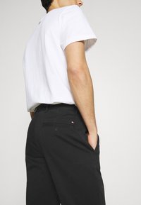 Homme portant une chemise blanche à manches courtes et un pantalon noir, main dans la poche, vu de dos et de côté sur un fond clair uni.