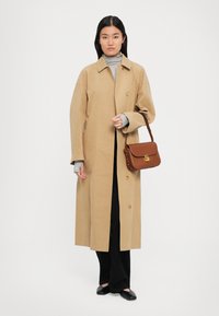 Kvinna som bär en beige lång kappa över en grå polotröja, svarta byxor, svarta skor och håller en brun axelväska mot en neutral bakgrund.
