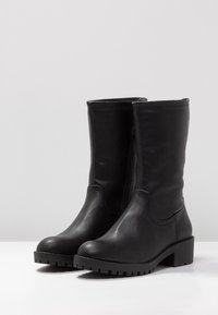 Botas de cuero negro hasta la rodilla con un acabado texturizado, punteras redondeadas, tacones cuadrados y una cremallera lateral para facilitar su uso.