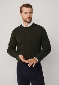 Maglione a trecce in lana verde scuro, scollatura tonda, polsini e orlo a costine, indossato sopra una camicia con colletto bianco, abbinato a pantaloni scuri.