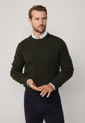 Dunkelgrüner Wollpullover im Kabelstrickmuster, Rundhalsausschnitt, gerippte Bündchen und Saum, über einem weißen Hemd mit Kragen getragen, kombiniert mit dunklen Hosen.
