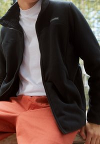 Giacca in pile nera con chiusura a zip e colletto alto, abbinata a una camicia bianca e pantaloni arancioni, con tessuto testurizzato e logo del marchio.