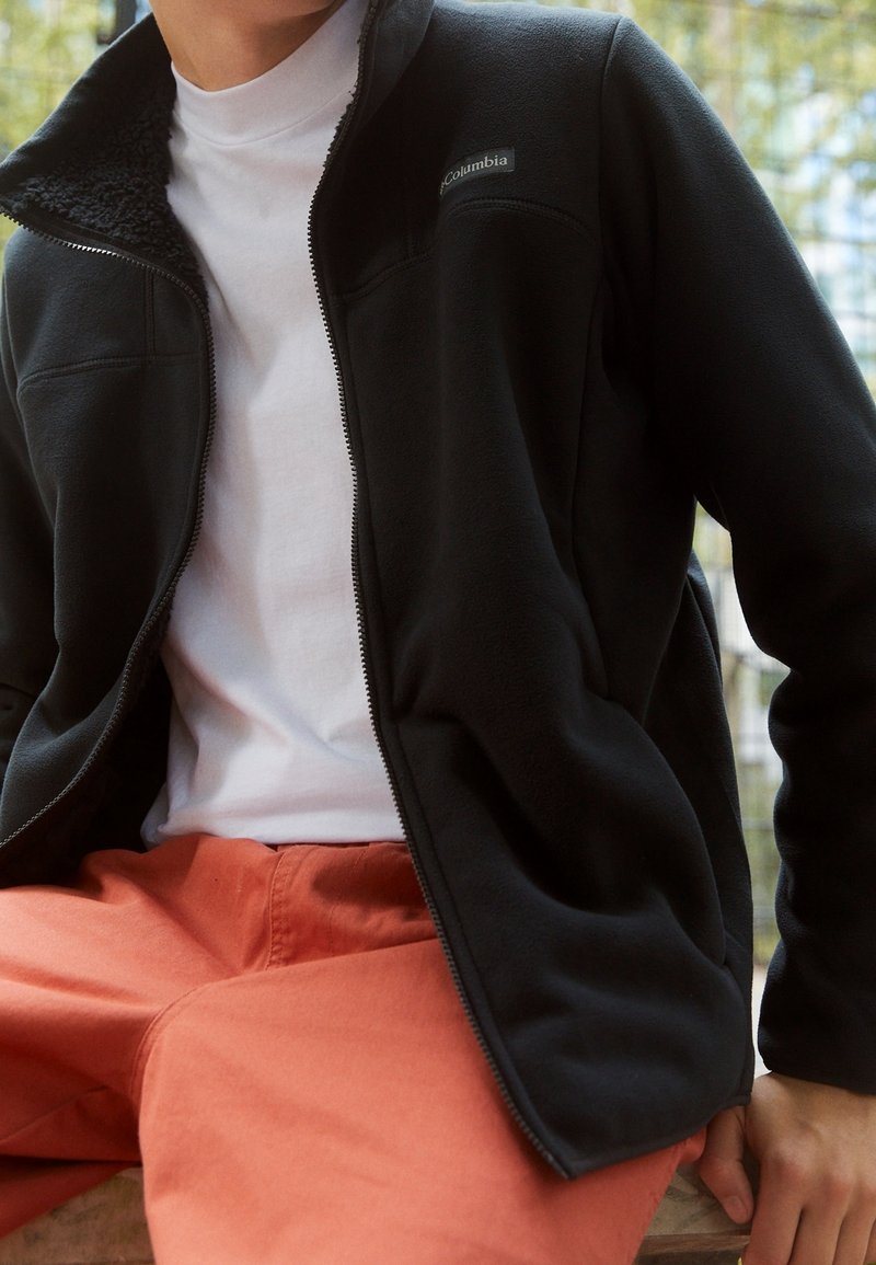 Giacca in pile nera con chiusura a zip e colletto alto, abbinata a una camicia bianca e pantaloni arancioni, con tessuto testurizzato e logo del marchio.
