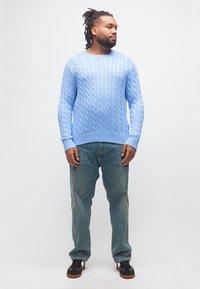 Άνδρας με πλεξούδες φοράει ανοιχτόμπλε πουλόβερ με πλεκτό σχέδιο, φαρδιά μπλε τζιν και μαύρα αθλητικά παπούτσια, στέκεται κοιτώντας στο πλάι.