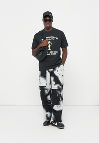 Svart grafisk t-shirt med vit text och tecknad bild, byxor i cargo-stil med svart och vit mönstring, svart keps, solglasögon och svarta sneakers.
