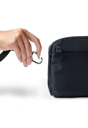 Hand holding a silver carabiner clasp, attaching it to the black zippered loop of a small fabric pouch or bag.
