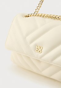 Kollane kreemjas käekott kuldse ketiga ja logoga detailidega. Omab diagonaalset õmblusmustrit ja siledat nahateksi.