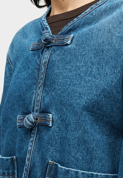 Τζιν μπουφάν σε μεσαίο μπλε χρώμα με υφή, διαθέτει κούμπωμα με κουμπιά κόμπου, δύο μπροστινές τσέπες και κλασικό γιακά.