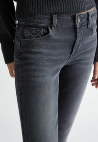 Vaqueros gris oscuro con corte ajustado, bolsillos frontales decorados con tachuelas y cierre de botón, confeccionados en un material de mezclilla suave.