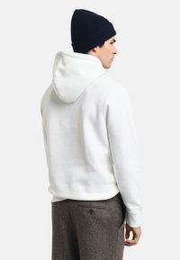 Sudadera blanca con capucha y textura suave, con un corte relajado y una gran capucha trasera. Combinada con pantalones gris oscuro y un gorro azul marino.